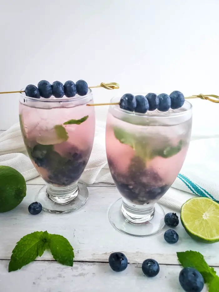 blueberry mojito in wine glass with fresh blueberries and mint, with sliced limes on wood backdrop